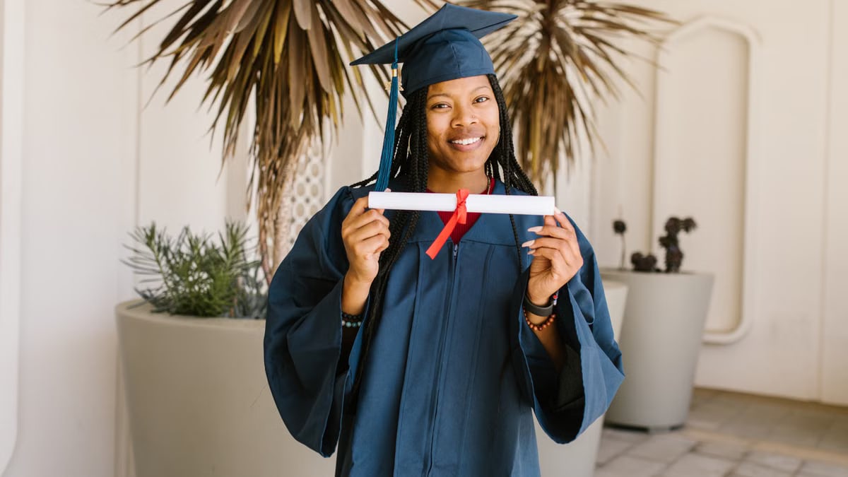 Young woman celebrating graduation with diploma in hand, wearing cap and gown, smiling proudly.